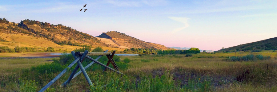 A buck and rail fence fills the foreground of a lush, green valley with layered rock formations in the background.