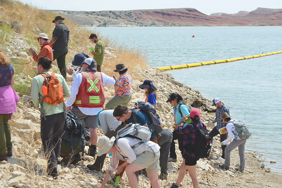 A group of geologists examine rocks along a lakeshore.