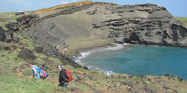 People walk on a grassy hillside with a dark sandy outcrop and a beach in the background.