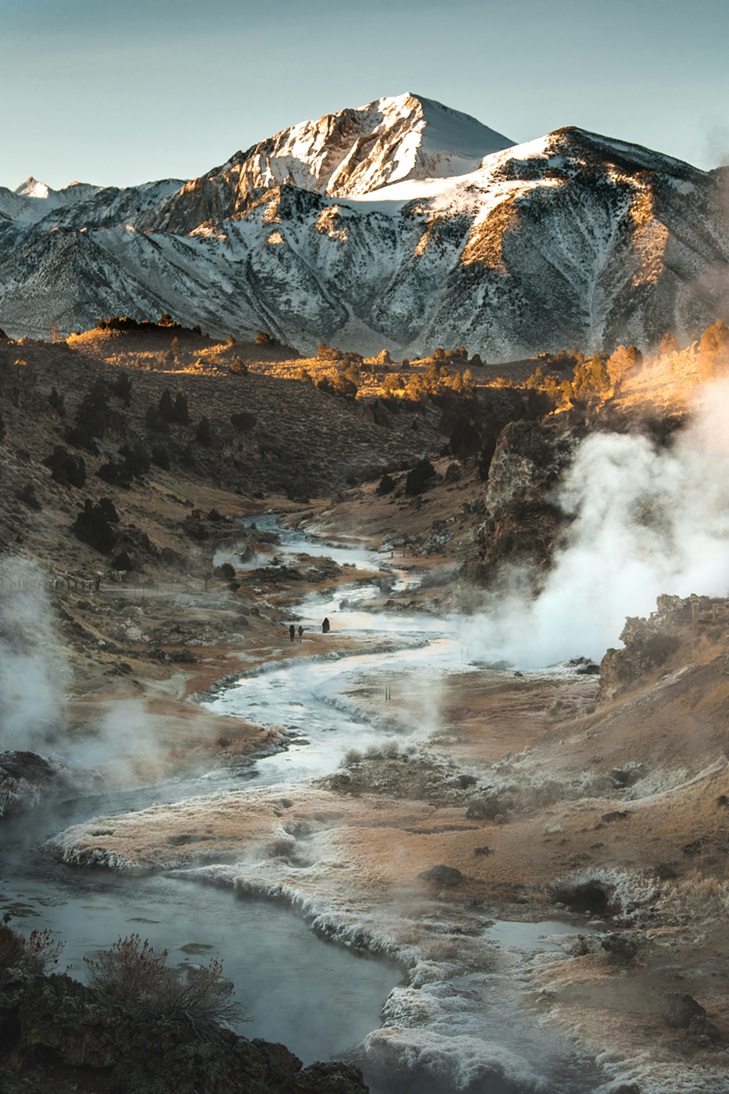 A snow-covered mountain lit up by the setting sun towers above a valley in which frost-covered, amber vegetation borders a snaking flow of water. Steam from the water rises into the air.