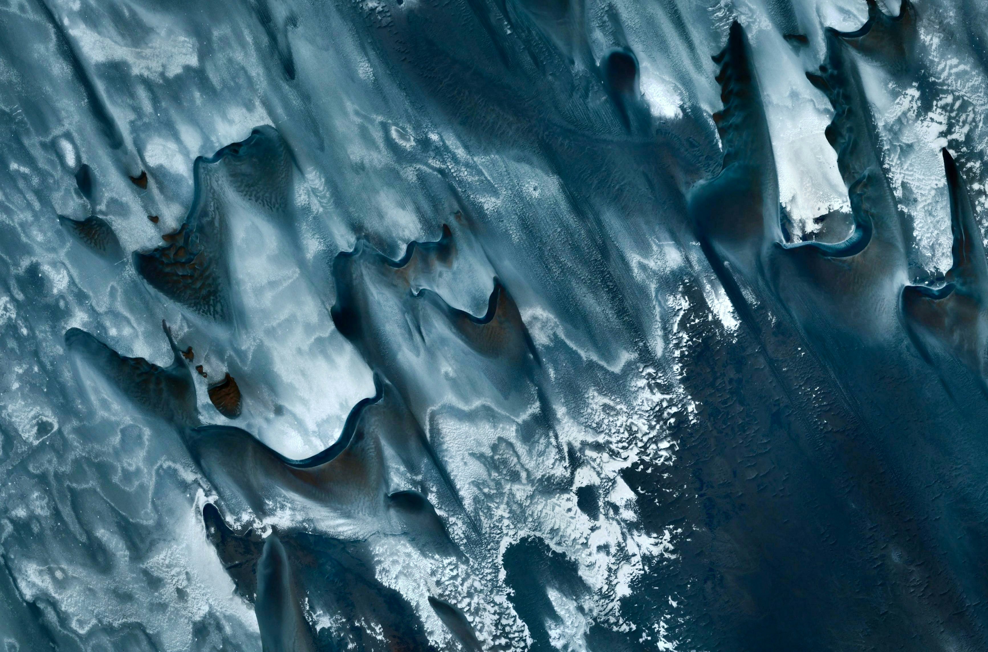 White and various tones of blue and gray make up this aerial photo of water, snow, and land or sand in this image captured from a plane.