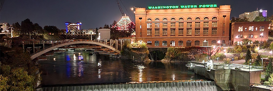 A nighttime shot of a river and the top of a small waterfall in front of a brick building.